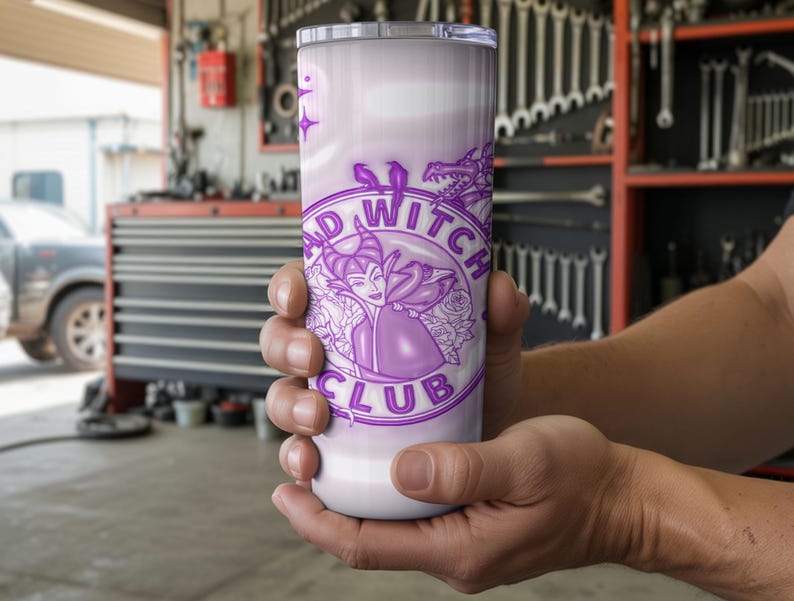 May include: A white and lavender tumbler featuring the text "BAD WITCH CLUB" in a circular design with a purple illustration of a witch. The tumbler is held in a person's hands. The background is a garage.