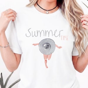 Puede incluir: Camiseta blanca con las palabras "Summer Time" en gris y rosa. La camiseta presenta una ilustración en acuarela de una persona con sombrero corriendo. El sombrero es gris y las piernas de la persona son rosas.