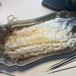 May include: A silver-toned, ornate tray holds a mass of white and cream-colored wool roving. Knitting needles and a crochet hook are arranged nearby. A small plant and scissors are also visible.