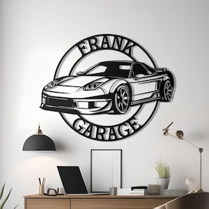 Op de afbeelding: Zwart metalen kunstwerk aan de muur met een autosilhouet en de tekst "FRANK GARAGE" in een cirkelvormig ontwerp. Het kunstwerk is bevestigd aan een witte muur boven een houten bureau.