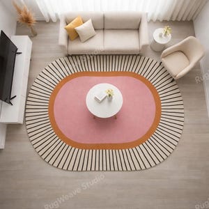 May include: An oval area rug featuring a central pink oval surrounded by a thick orange border and an outer cream-colored section with radiating black lines. The rug is positioned on a light wood floor in a room with a sofa, armchair, and table.