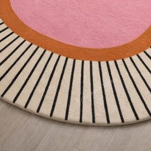 May include: A circular rug featuring a large central pink circle surrounded by a thick orange border. The outer edge of the rug is cream-colored and decorated with a series of evenly spaced, thin black lines radiating outward from the center.