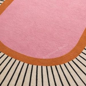 May include: A tufted area rug featuring a large central pink oval shape bordered by a thick burnt orange band. The background consists of a cream-colored field with radiating black lines extending outward. The text RugWeave Studio is visible.
