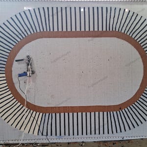 May include: A large oval rug in progress on a white backing frame, featuring a thick brown border and a series of radiating black yarn lines. A blue and silver tufting gun is attached to the left side, with white yarn trailing from it.