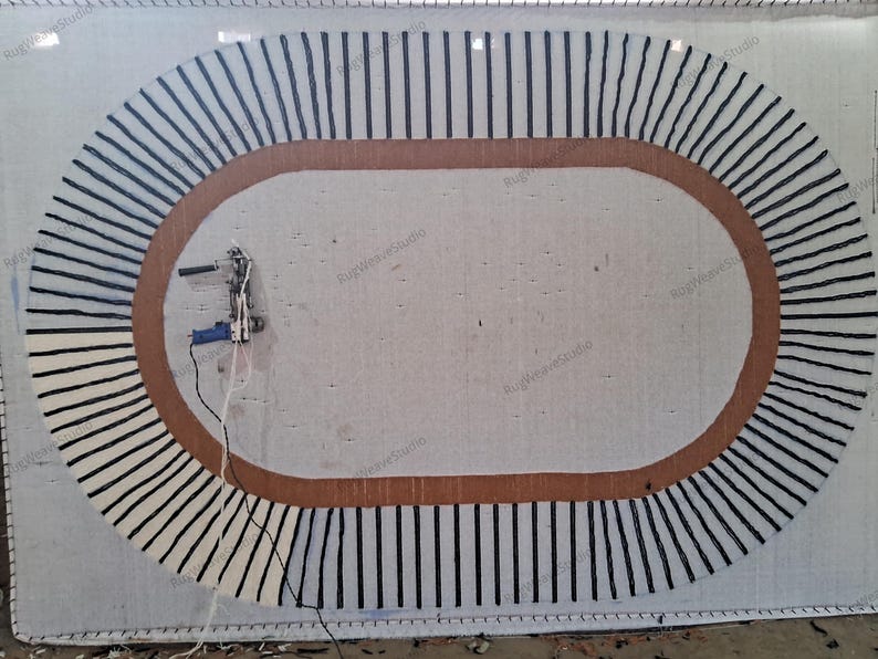 May include: A large oval rug in progress on a white backing frame, featuring a thick brown border and a series of radiating black yarn lines. A blue and silver tufting gun is attached to the left side, with white yarn trailing from it.