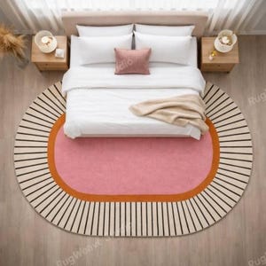 May include: An oval area rug featuring a central pink oval surrounded by an orange border and an outer cream-colored section with radiating black lines. The rug is positioned beneath a bed with white bedding and a small pink accent pillow.