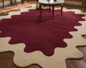 Op de afbeelding: Een groot, uniek gevormd vloerkleed met een bordeauxrood midden en een crèmekleurige, golvende rand. Het vloerkleed ligt op een houten vloer, en een kleine tafel en stoelen zijn zichtbaar op de achtergrond. De afmetingen van het vloerkleed zijn niet zichtbaar.