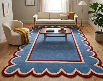 May include: A large rectangular area rug featuring a solid blue center, a cream-colored inner border, and a scalloped edge outlined in deep red. The rug has a flat-woven texture and covers a significant portion of the light-colored wooden floor.