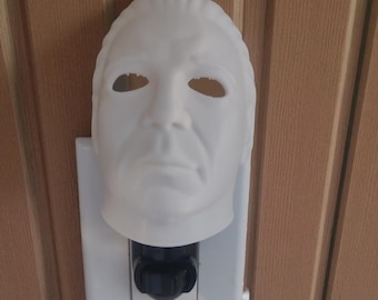 May include: A white plastic mask resembling a character from a horror film, mounted above a white electrical outlet. The mask has eye and mouth openings and is attached to the outlet with a black plug inserted. The background is a brown wooden surface.