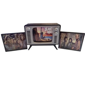 May include: A Planet of the Apes themed decorative set featuring a retro-style television with the movie title on the screen, flanked by two framed images of characters from the film. The TV is black and white with a silver trim.