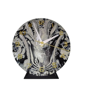 May include: Round clock featuring a black and white alien design with gold-colored numerals and hands. The clock has a black base and is a decorative item.