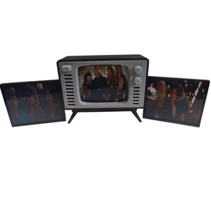 May include: A retro-style television set with a black and silver frame, featuring a screen displaying a group of people. Two additional panels on either side show similar images. The TV has a vintage aesthetic.