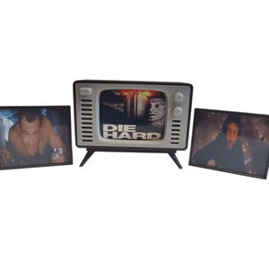 Könnte beinhalten: Ein Fernseher im Retro-Stil mit dem Schriftzug "DIE HARD" auf dem Bildschirm, flankiert von zwei gerahmten Bildern. Der zentrale Fernseher hat einen schwarzen Rahmen, eine weiße Front und schwarze Beine. Die Bilder zeigen Szenen aus dem Film.