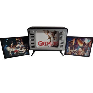 Op de afbeelding: Een retro-stijl televisie met het woord "Gremlins" in het rood op het scherm. Twee ingelijste afbeeldingen met scènes uit de film staan aan weerszijden. De tv is zwart en grijs met een vintage design.