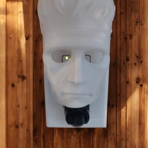 May include: A white, wall-mounted soap dispenser shaped like Frankenstein's monster's head. The dispenser has a black nozzle and is mounted on a white rectangular base. The background is a wooden wall.