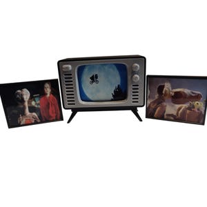 May include: A miniature retro television with a black frame and a screen displaying a silhouette of a bicycle flying in front of a full moon. Two framed images of the movie E.T. are on either side of the TV.