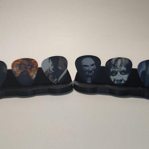 May include: A black guitar pick holder with six guitar picks featuring horror movie characters. The picks are arranged in two rows of three. The characters on the picks include Ghostface, Leatherface, Jigsaw, Freddy Krueger, Jason Voorhees, and Michael Myers.