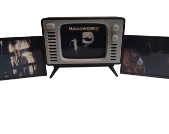 May include: A retro-style television with a black and white screen displaying the movie "Halloween 5". The TV has a white and black frame with two side panels. The TV has a black stand.