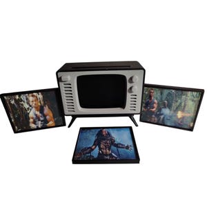 Peut inclure: Une télévision miniature noire et blanche avec trois images encadrées. Les images présentent des scènes et des personnages du film "Predator". La télévision a un design rétro avec un écran noir et des accents blancs.