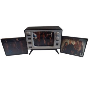 May include: A miniature black and white television with a screen displaying a movie scene. Two additional framed images flank the TV, also showing movie stills. The TV has a retro design with knobs and a small screen.