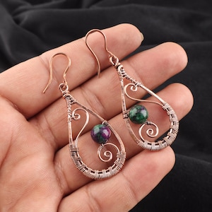 May include: Copper wire wrapped earrings with a teardrop design. Each earring features a green and purple gemstone bead. The earrings have a hook closure and a decorative swirl design.