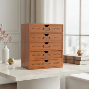 Pode incluir: Um baú de armazenamento de madeira com cinco gavetas castanhas claras. Cada gaveta tem as palavras "MERCERIE" e "BOUTON" em preto. O baú tem porta-etiquetas de metal e está sobre uma mesa de mármore branco. Altura aproximada: 30 cm.
