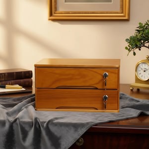 May include: A two-drawer wooden storage box with a warm, natural finish. Each drawer has a key lock and a small, recessed handle. The box is set on a dark wooden surface, with a clock and books in the background.