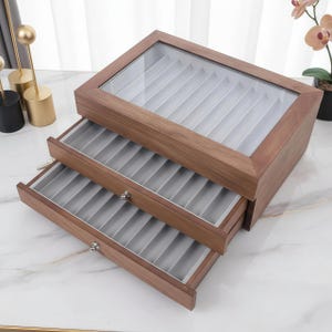 May include: A wooden jewelry box with a glass top and two pull-out drawers. The box is made of dark brown wood and has a light gray interior. The drawers have small silver knobs. The top of the box is clear, allowing a view of the contents.