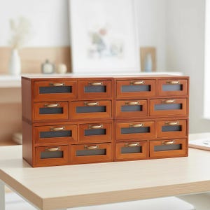 Puede incluir: Un mueble de almacenamiento de madera con dieciséis cajones pequeños. El mueble está hecho de madera marrón cálida y presenta manijas de color latón. Cada cajón tiene una pequeña ventana rectangular. El mueble está sobre una mesa de color claro.
