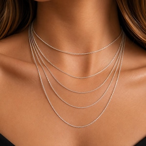 Op de afbeelding: Een set van vijf gelaagde zilverkleurige kabelkettingen van verschillende lengtes. Elke ketting heeft een delicaat, uniform schakelontwerp met een gepolijste metalen afwerking. De kettingen zijn in een trapsgewijze formatie rond de hals geplaatst.