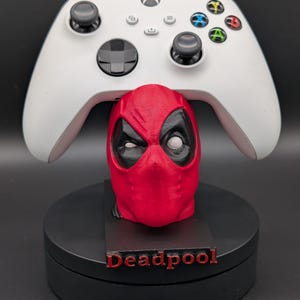 May include: A white Xbox controller rests on a red Deadpool head stand. The stand is on a black base with the name "Deadpool" in red. The controller has black joysticks and colored buttons.