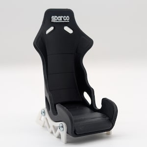 May include: A black Sparco racing seat model with a white base. The seat has a high back with the Sparco logo in white. The model is a miniature replica of a racing car seat.