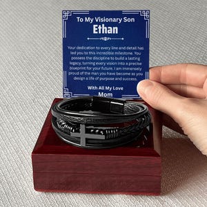 May include: A black leather bracelet with a silver cross charm, presented in a dark red wooden box. A card with the text "To My Visionary Son Ethan" is held above the bracelet.
