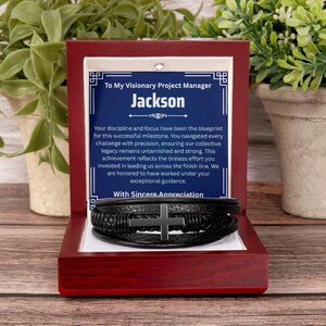May include: A black multi-strand leather bracelet with a central matte black cross charm, presented in a polished dark wood gift box. The box interior features a blue card with white text addressed to Jackson, expressing appreciation for a project manager.