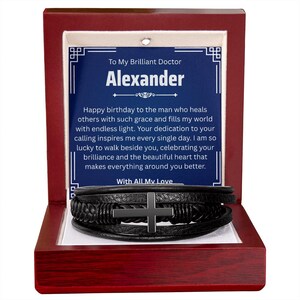 May include: A black leather bracelet with a silver cross is displayed inside a red wooden box. The box contains a blue card with the text "To My Brilliant Doctor Alexander" and a birthday message.