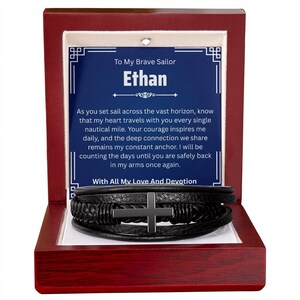 May include: A black leather bracelet with a silver cross is displayed inside a wooden box. The box contains a blue card with the text "To My Brave Sailor Ethan" and a heartfelt message. The bracelet is a multi-strand design.