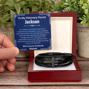 May include: A black leather multi-strand bracelet featuring a central metal cross charm, presented inside a dark wood box with a white interior. A blue card with white text reads: To My Visionary Florist Jackson, With Sincere Gratitude Mateo.