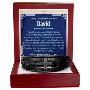 May include: A black leather bracelet with a silver cross is displayed inside a wooden box. The box has a red exterior and a white interior. A card inside the box has text that reads "To My Compassionate Son David".