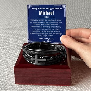 May include: A black leather bracelet with a silver cross charm, presented in a dark red wooden box. A card with the text "To My Hardworking Husband Michael" is held above the bracelet.