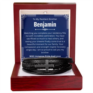 May include: A black leather bracelet with a silver cross is displayed inside a red wooden box. The box contains a blue card with the text "To My Resilient Brother Benjamin" and a heartfelt message. The bracelet is a multi-strand design.