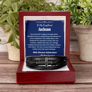 May include: A black multi-strand leather bracelet featuring a central metal cross charm, presented in a dark wood gift box. The box contains a blue card with the text: To My Exceptional Jackson, You have built a legacy through sheer discipline.