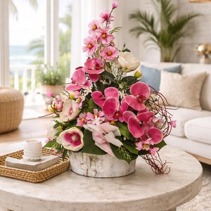 May include: A colourful floral display with pink orchids and other flowers in a distressed white wooden container. The arrangement is on a round, light-coloured coffee table, creating a focal point in a bright living room setting.