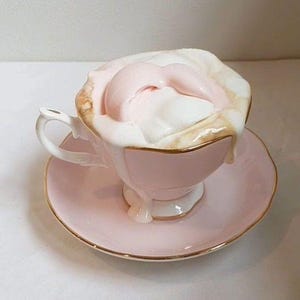 Könnte beinhalten: Eine rosa Teetasse und Untertasse mit Goldrand, überfüllt mit einem weißen und rosa Dessert. Die Tasse hat einen zarten Henkel und steht auf einer passenden Untertasse. Das Dessert scheint ein geschichtetes Konfekt zu sein.