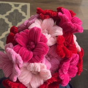 May include: A handmade flower bouquet in red, pink, and light pink. The flowers are made of a soft, textured material, and are arranged in a round shape. A craft item, the bouquet is a vibrant display of colour.