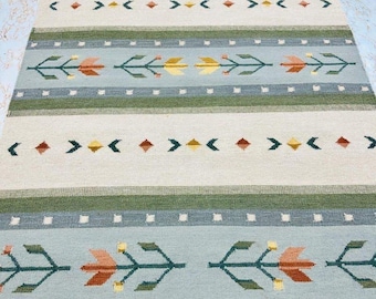 May include: A rectangular area rug featuring horizontal bands of light blue, cream, and dark green. The design includes repeating patterns of stylized floral motifs in orange and yellow, interspersed with rows of small white squares and geometric shapes.