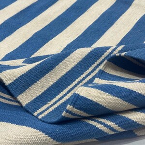 May include: A blue and white striped rug with a folded corner. The rug features alternating wide stripes of blue and off-white. The fabric appears to be woven, with a textured surface.