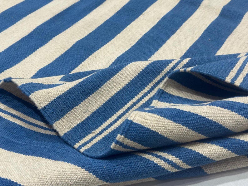 May include: A blue and white striped rug with a folded corner. The rug features alternating wide stripes of blue and off-white. The fabric appears to be woven, with a textured surface.