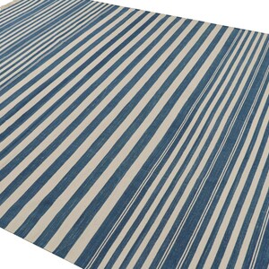 May include: A large, rectangular rug with a repeating pattern of blue and off-white stripes. The stripes run diagonally across the rug, creating a visual texture. The rug has a fringed edge on one side.