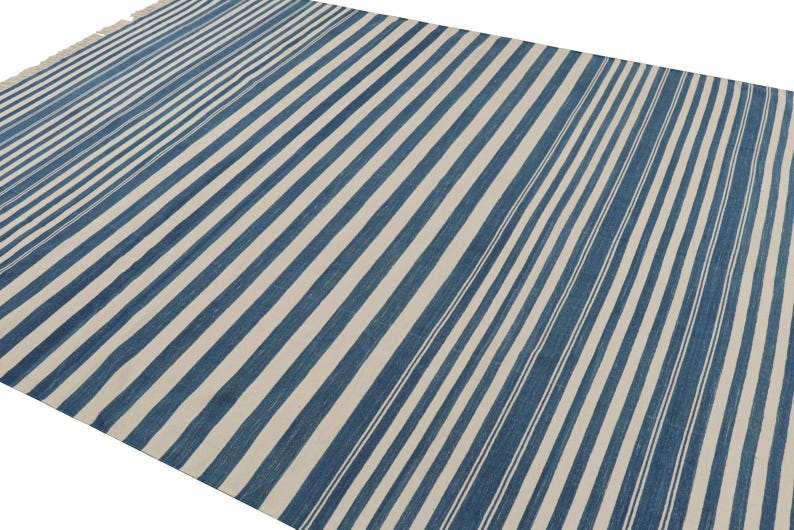 May include: A large, rectangular rug with a repeating pattern of blue and off-white stripes. The stripes run diagonally across the rug, creating a visual texture. The rug has a fringed edge on one side.