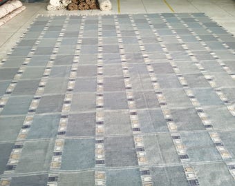 May include: A large, rectangular rug with a repeating pattern of rectangular blocks in shades of blue and gray. The blocks are separated by vertical stripes with a mix of white, beige, and dark blue accents. The rug is made of a woven material.